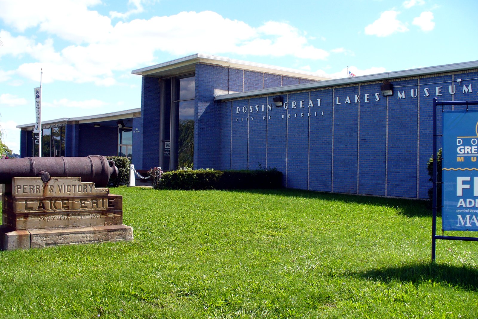 A low blue museum building with a sign reading Dossin Great Lakes Museum, a green lawn and a stone marker in front.