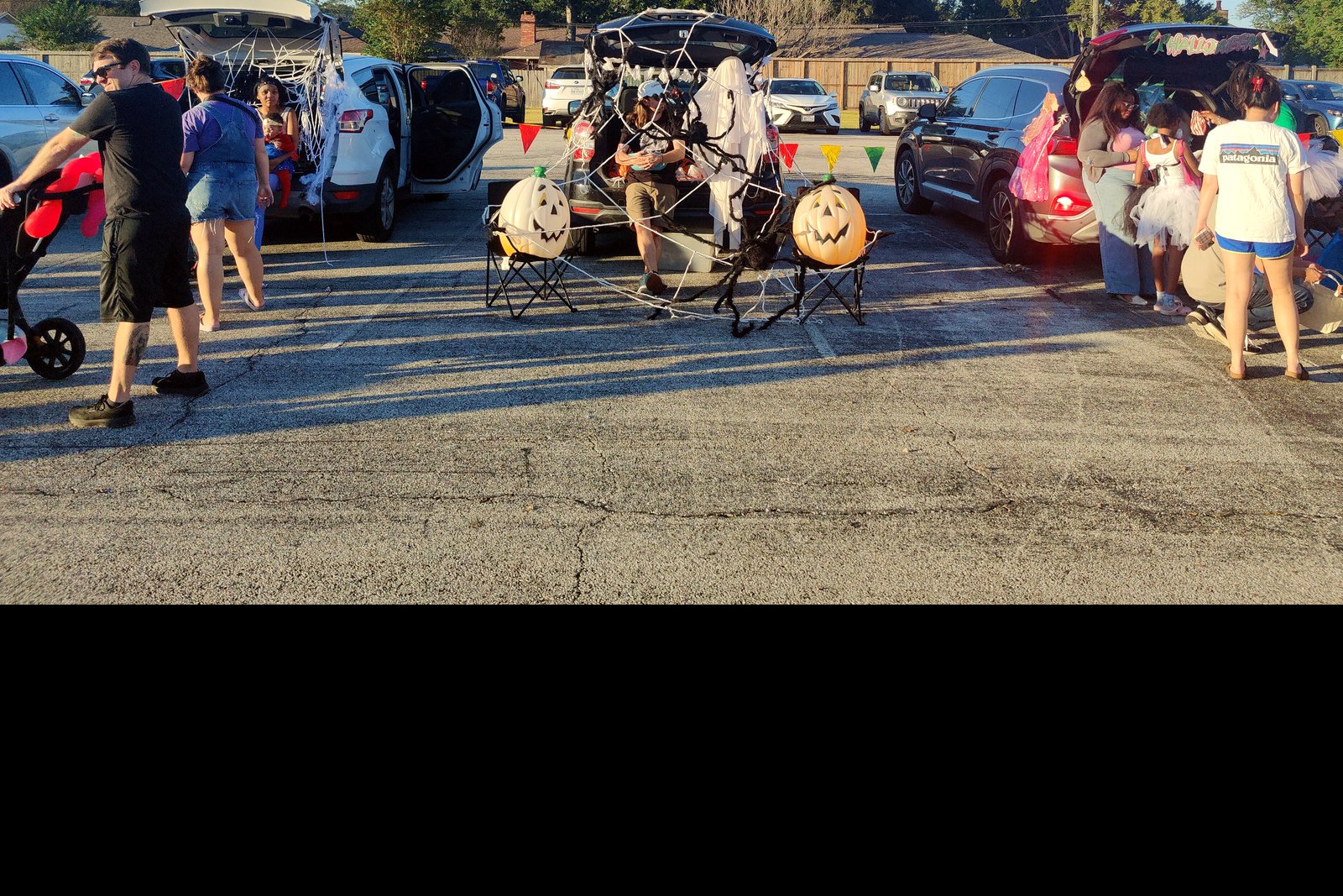 Families with children in costumes walk among parked cars with open trunks decorated for Halloween in a church parking lot on a sunny afternoon.