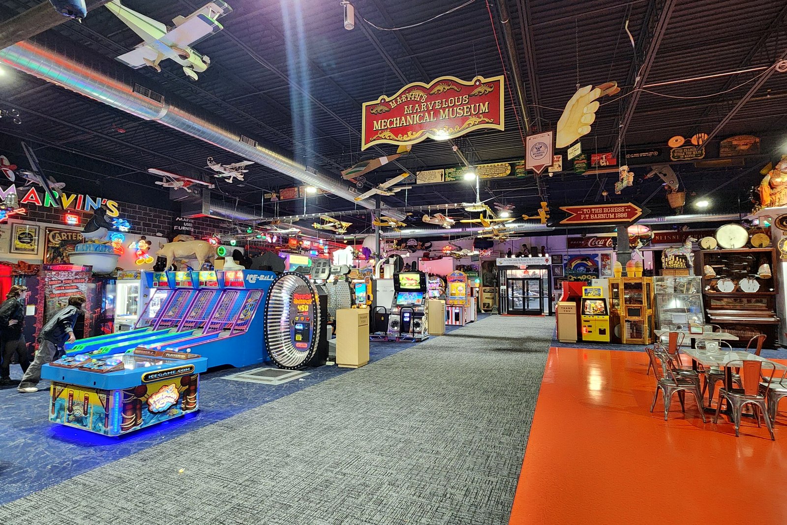 The brightly lit interior of Marvin’s Marvelous Mechanical Museum, with arcade games, model airplanes hanging from the ceiling and an orange floor.