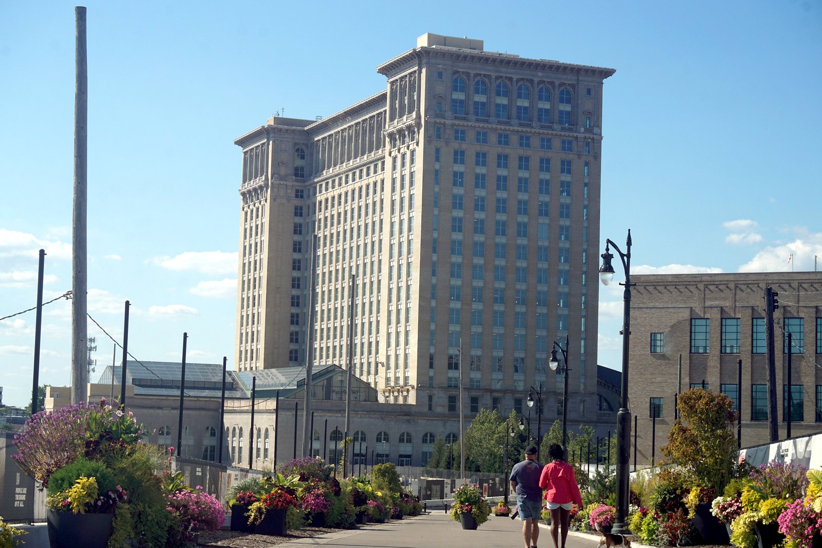 A tall beige train station tower behind a sidewalk plaza with flower planters, under a blue sky.