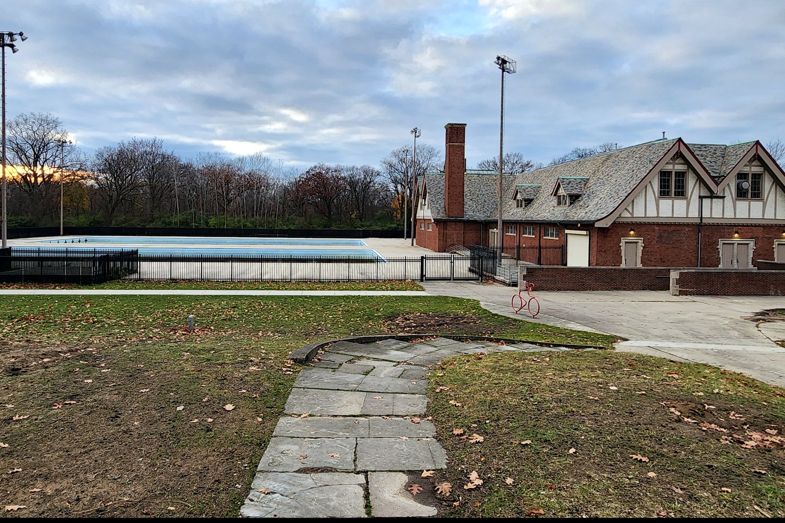 An empty outdoor swimming pool behind a black fence beside a brick Tudor-style bathhouse, with bare autumn trees under a cloudy sky.