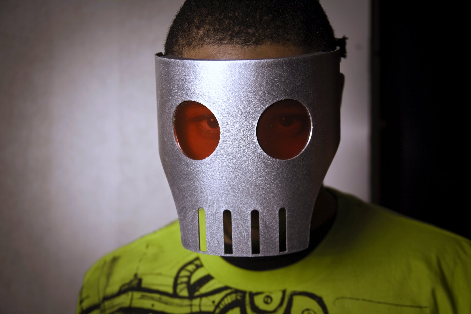 A person in a silver skull-like mask with round red eye holes and a bright green T-shirt stands against a plain gray wall.