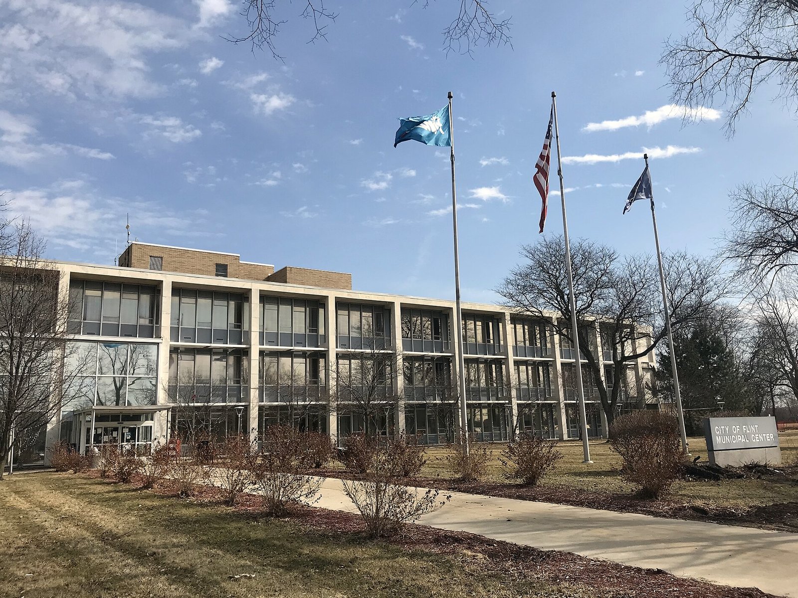 The Flint Municipal Center, a long four-story building of pale concrete and dark glass, behind a lawn with three flagpoles and a stone sign reading City of Flint Municipal Center, under a blue sky with bare trees.