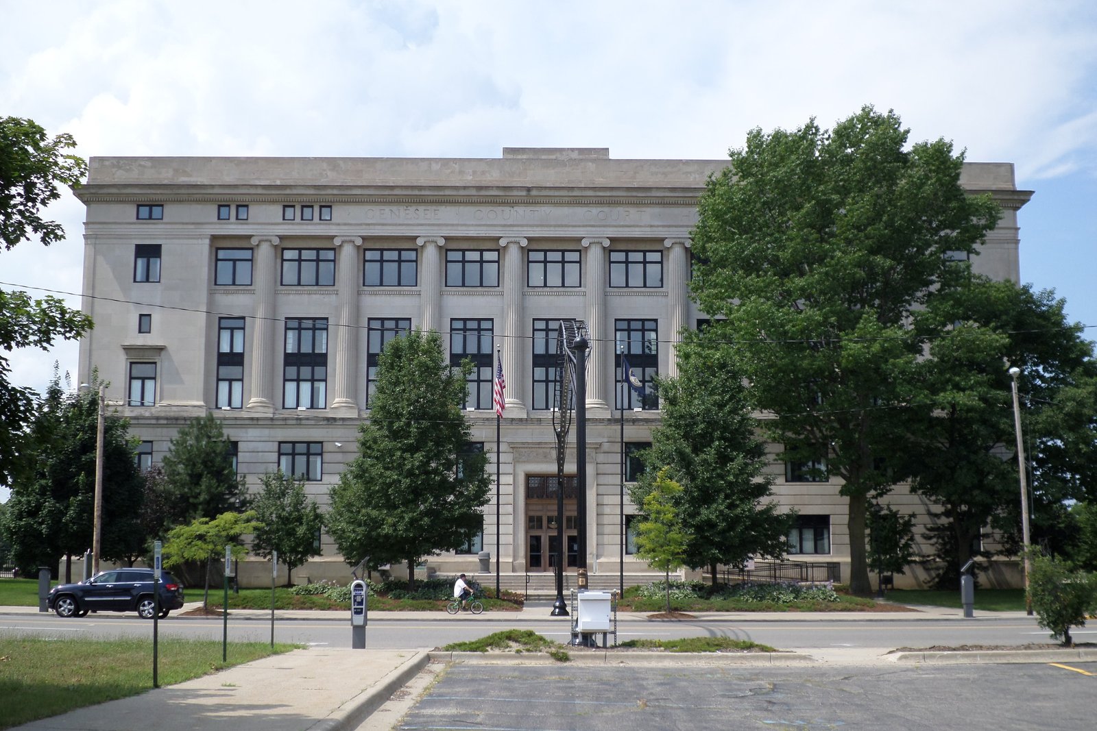 A four-story limestone courthouse with tall columns, an American flag and mature trees in front, seen from across a street on a summer day.