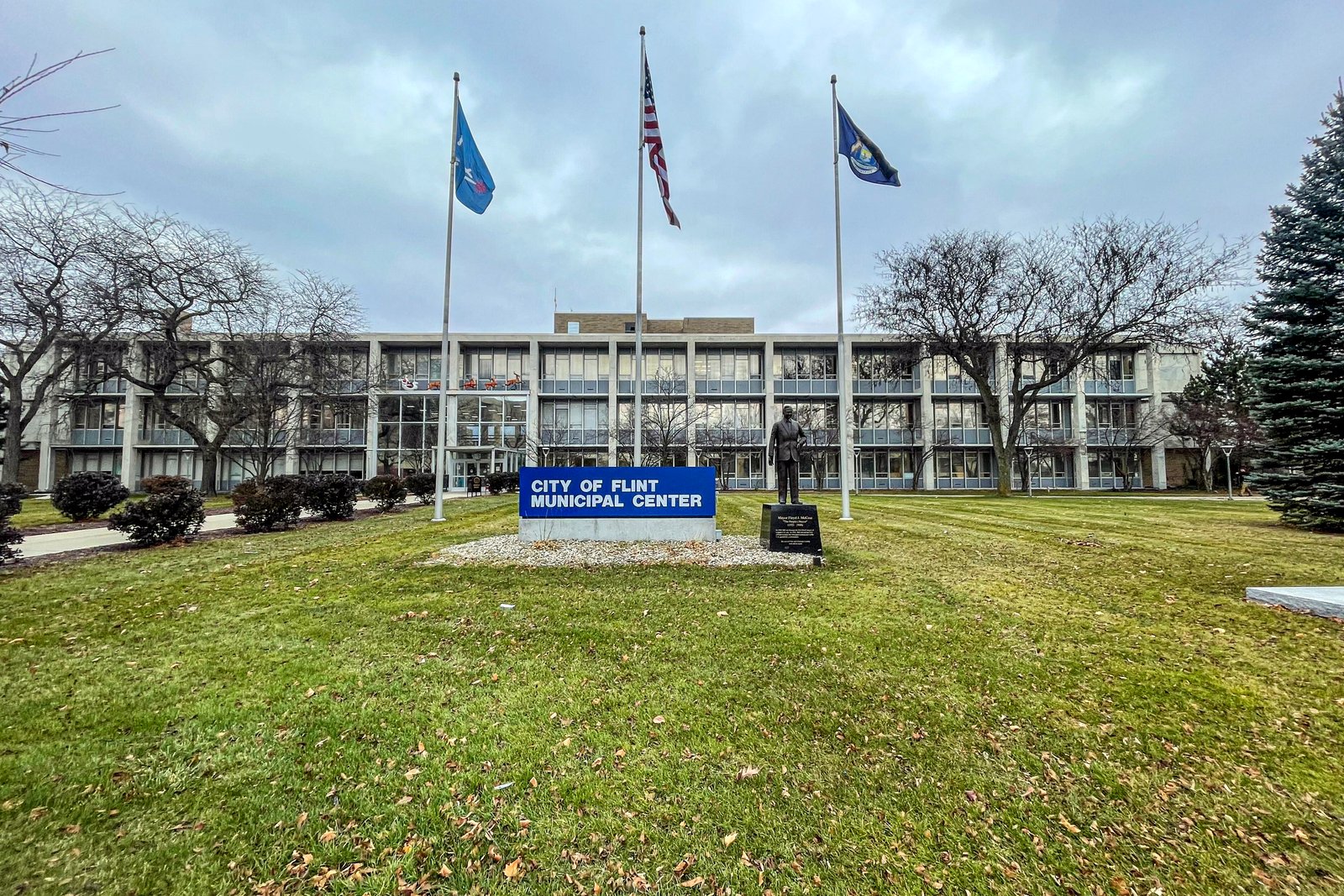 The City of Flint Municipal Center, a long low-rise office building behind a lawn with three flagpoles and a blue sign, under a cloudy sky.
