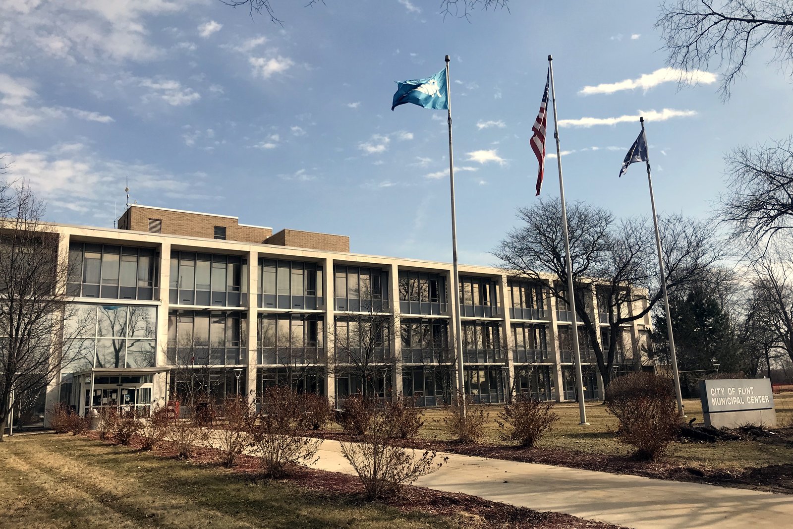 A four-story office building with long rows of windows behind three flagpoles, one flying the U.S. flag, with a lawn sign reading City of Flint Municipal Center.