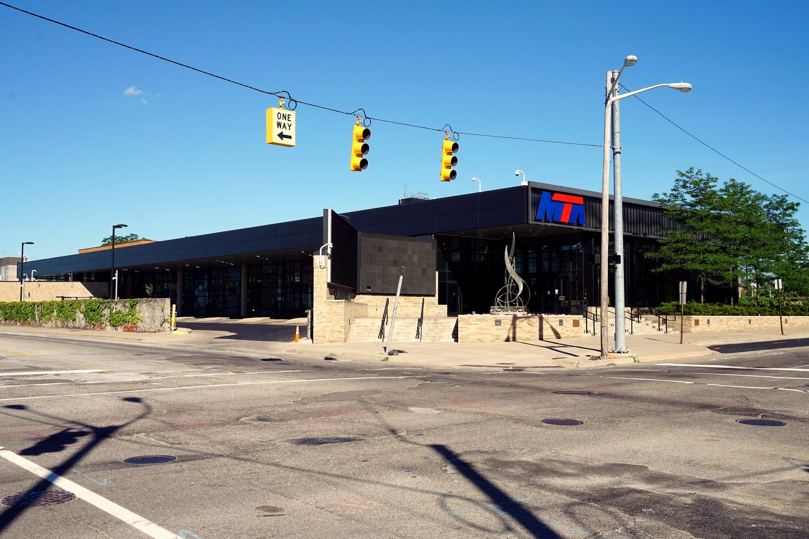 The MTA transit center in downtown Flint, a long, low dark building with the MTA logo, seen across an empty intersection with traffic lights under a blue sky.