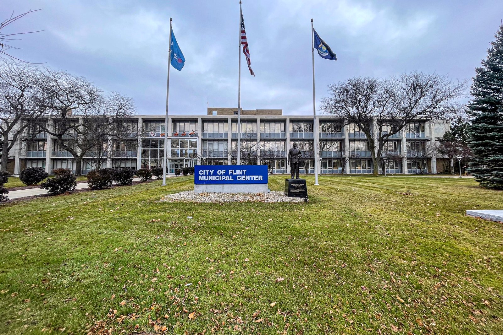 A long, low municipal office building behind a lawn with three flagpoles and a blue sign reading City of Flint Municipal Center.