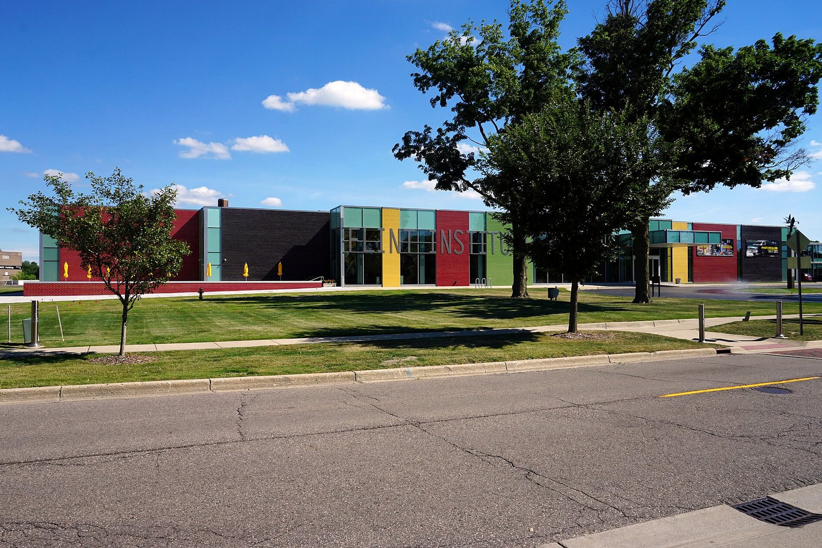 A long, low museum building with panels of red, black, green, yellow and teal brick behind a wide lawn and two tall shade trees, under a blue sky with scattered clouds.