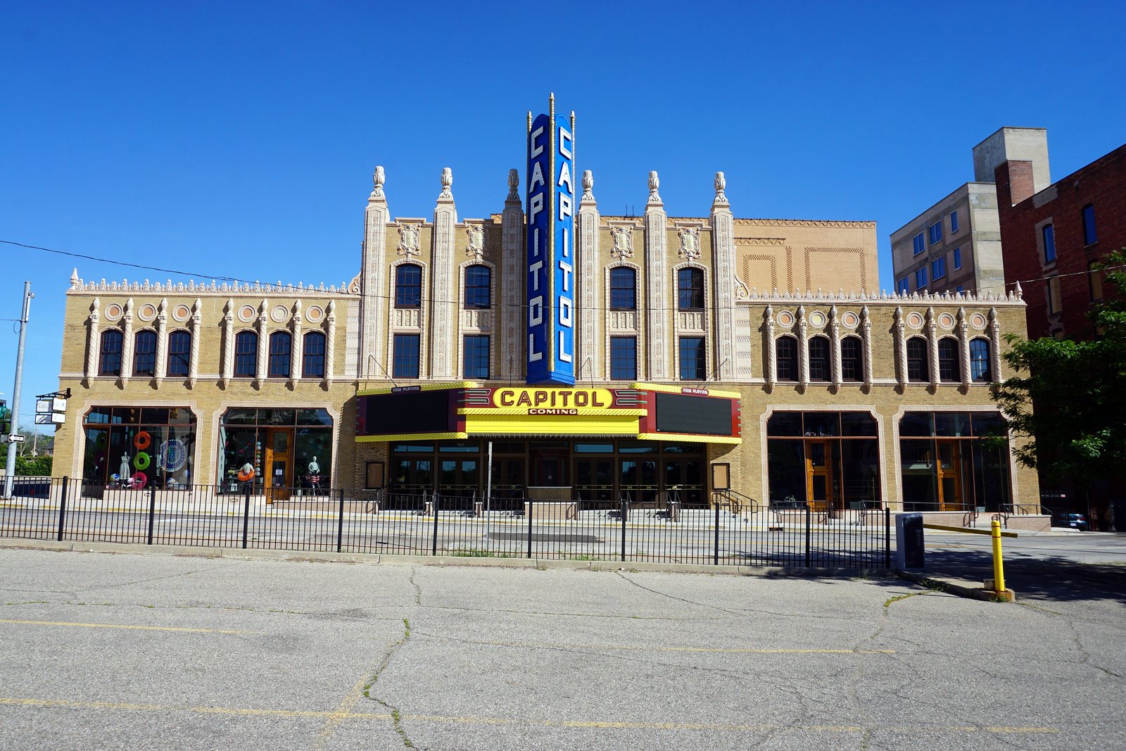 The tan brick Capitol Theatre in downtown Flint, with its tall blue vertical Capitol sign and marquee, under a clear blue sky.