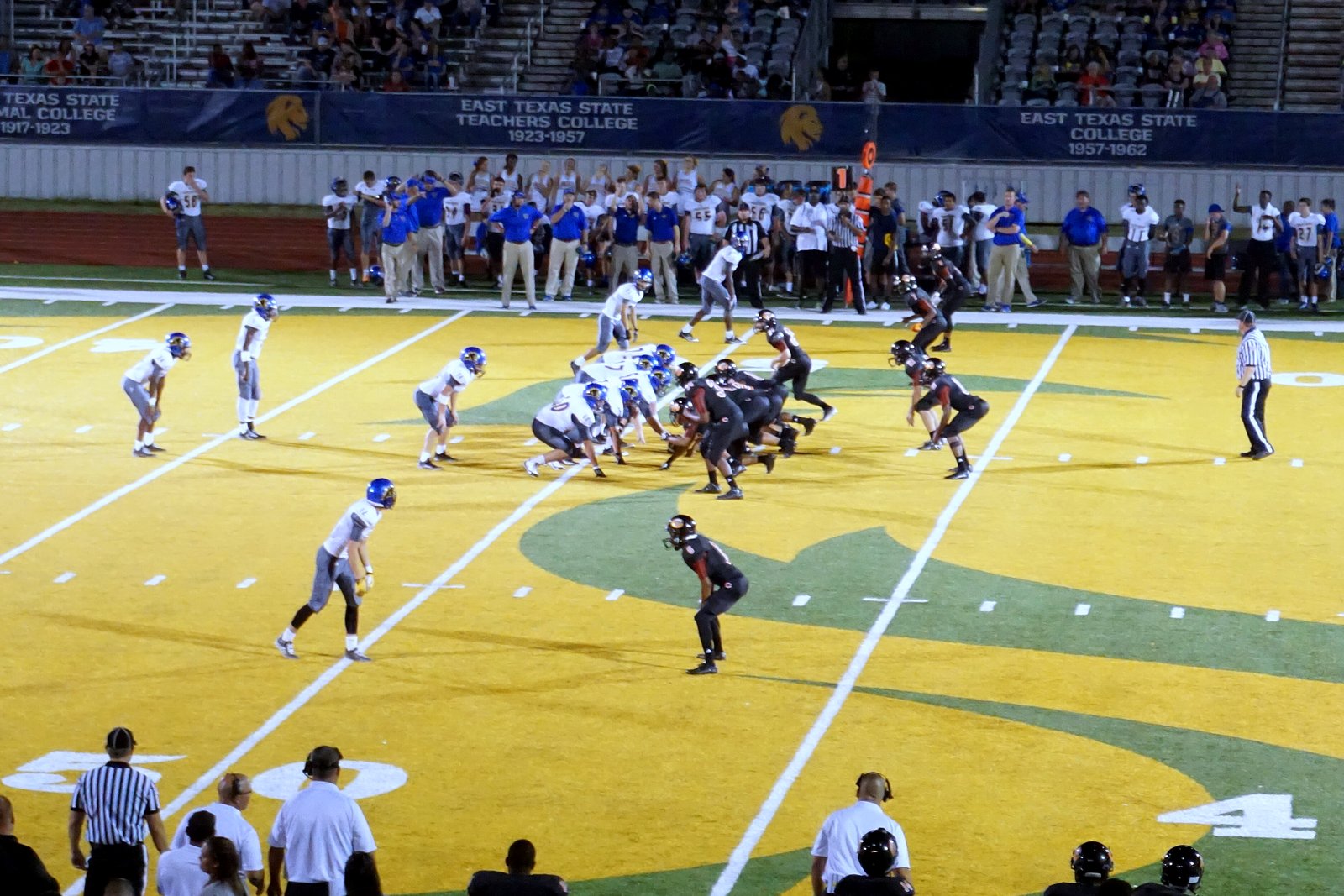 High school football players run a play on a field with a yellow end zone and a painted midfield logo, with coaches and players along the sideline in front.