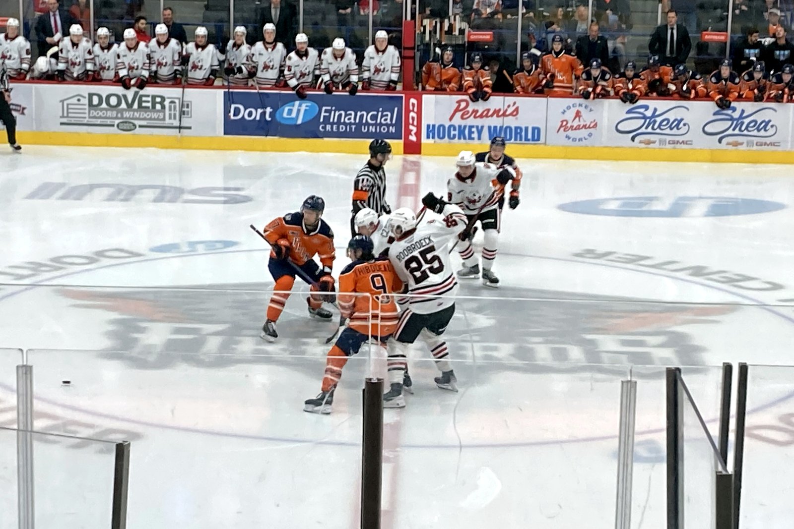 Hockey players in orange and in white jerseys line up for a faceoff at center ice inside a Flint arena, with players on the benches and fans behind the boards.