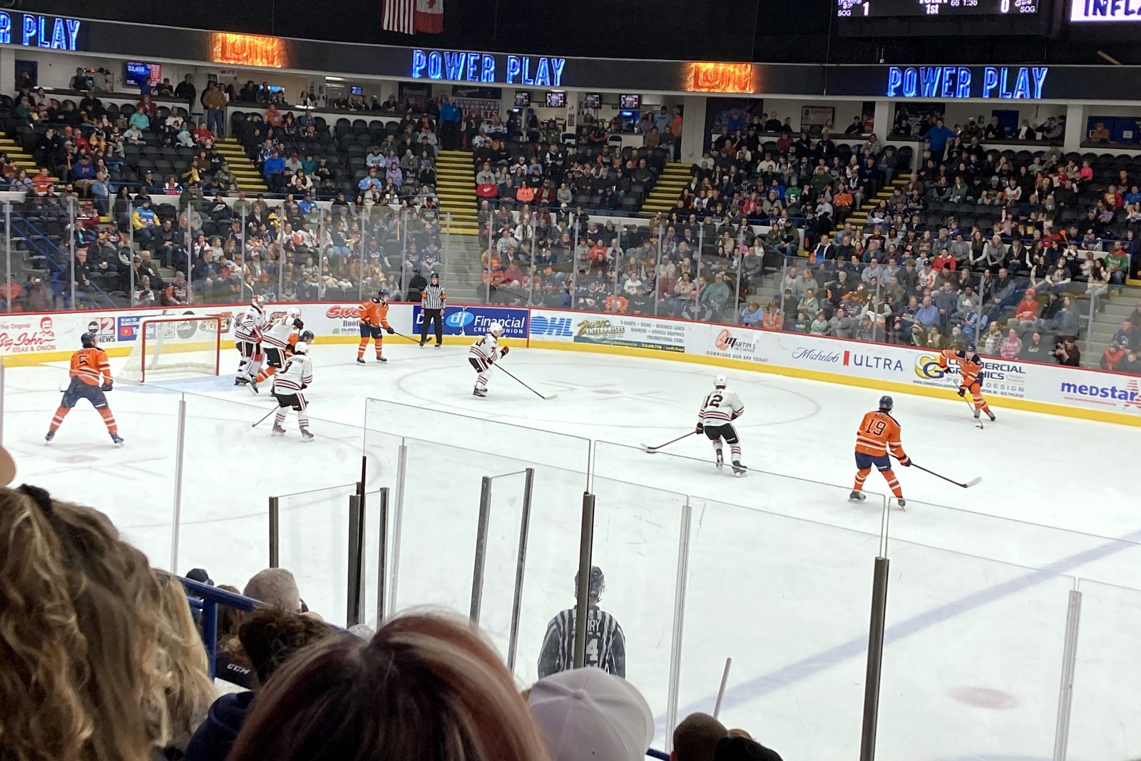 Hockey players skate on the ice in front of a crowded arena with blue signs reading Power Play along the upper deck.