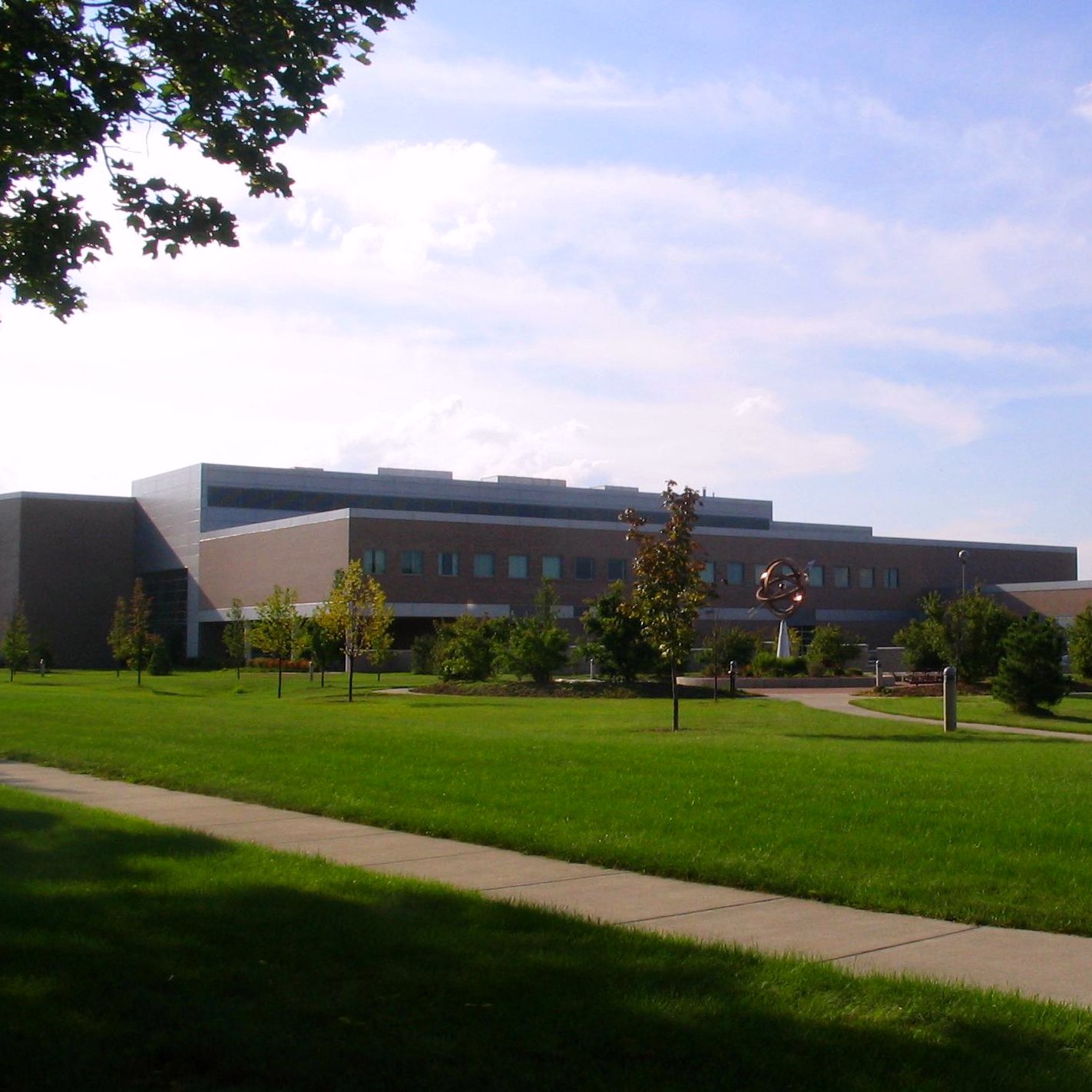A long, low modern brick and glass college building beyond a wide green lawn and a curving sidewalk, with young trees and a tall leafy tree framing the left side under a blue sky.