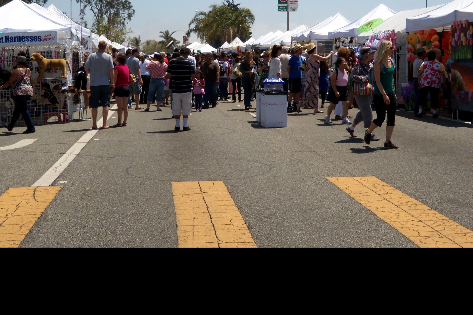 A crowd of shoppers walks down a closed street between rows of white vendor tents on a sunny day, with palm trees in the background.