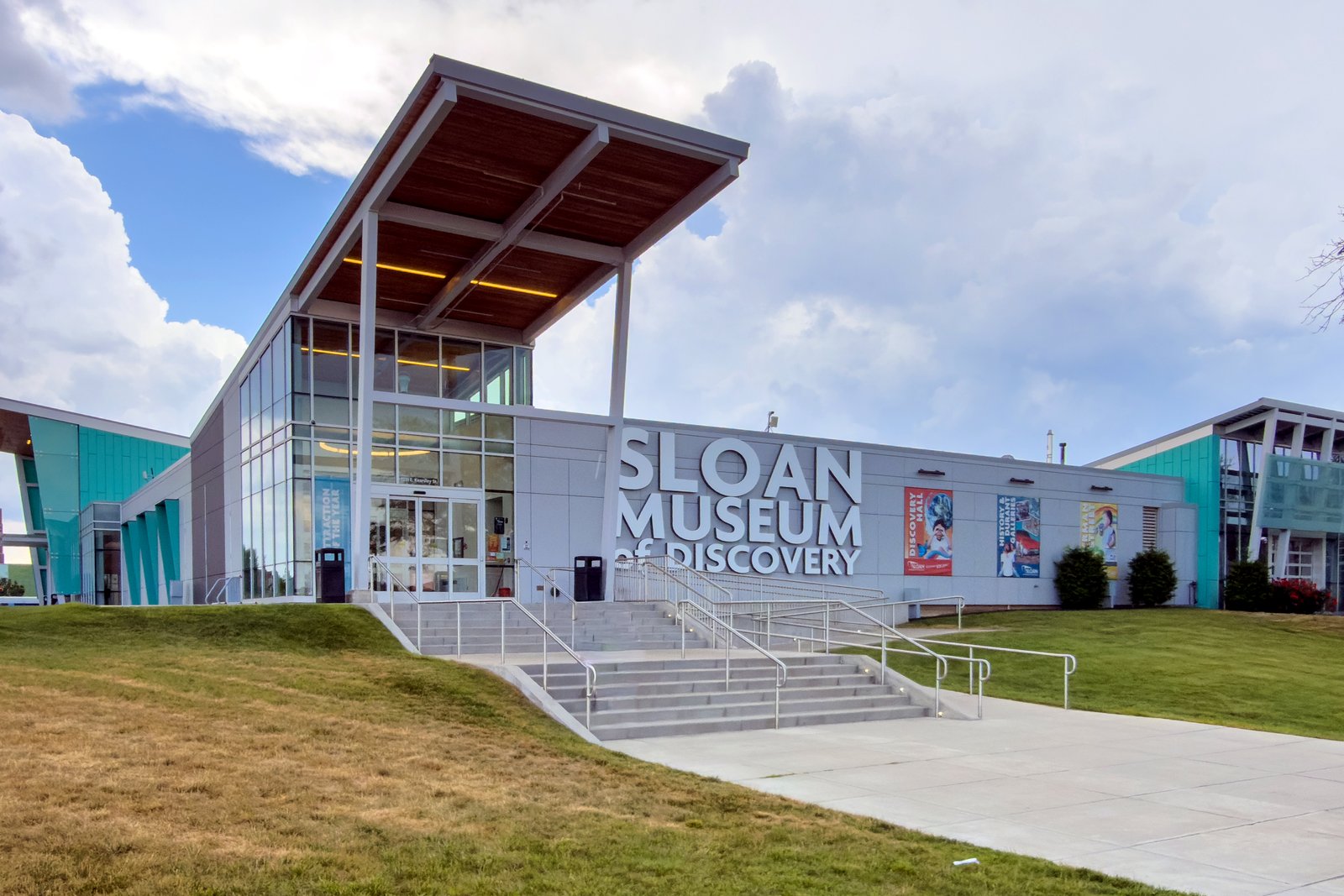 The glass-fronted entrance of the Sloan Museum of Discovery in Flint, with a wooden overhang, silver lettering and colorful exhibit banners beside a walkway and lawn.