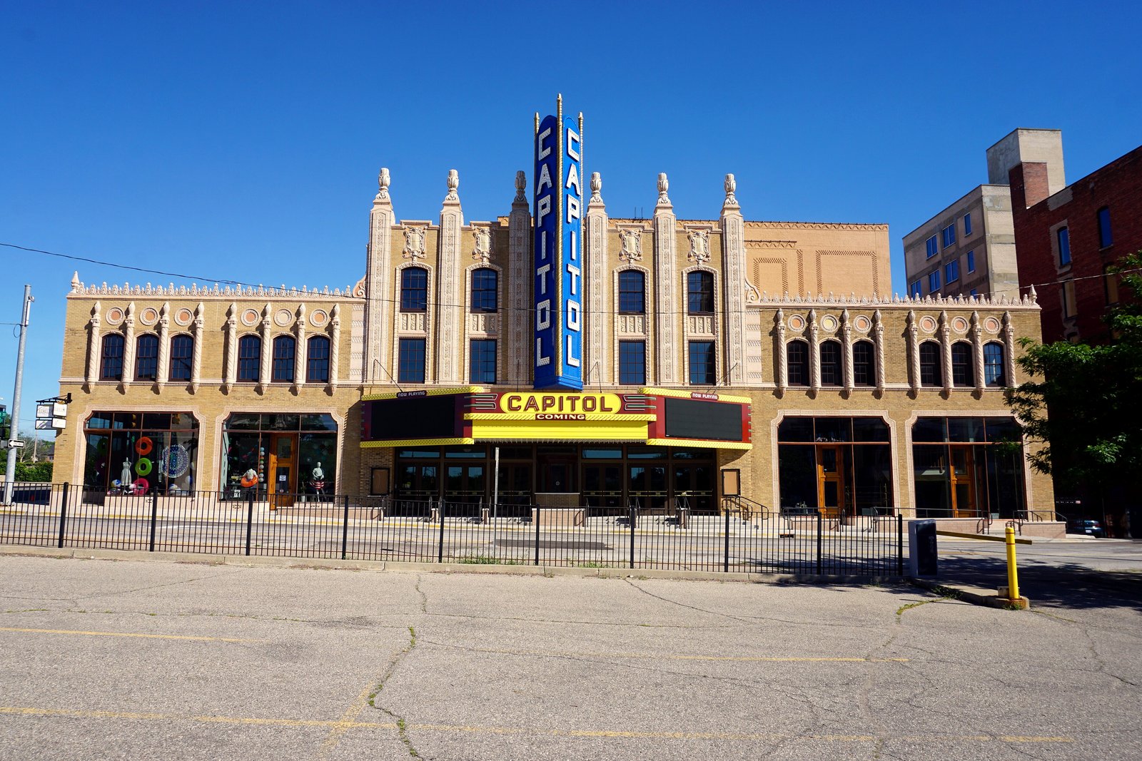 The yellow marquee and tall blue vertical Capitol sign on the tan facade of Flint’s Capitol Theatre under a clear blue sky.