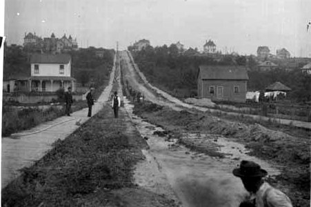 Workers lay a large water pipe in an open street trench in a historic black-and-white construction photo.