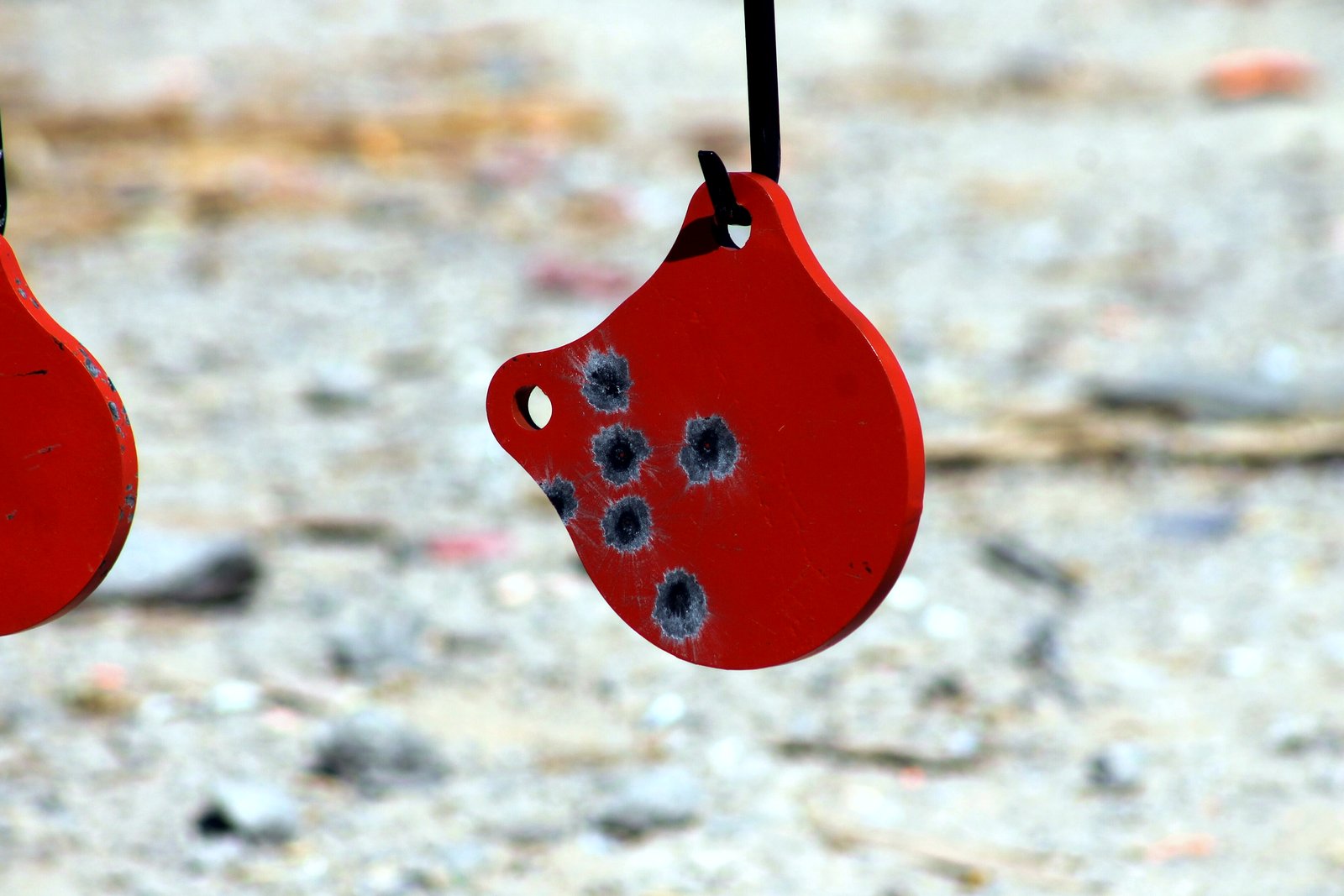 A red steel shooting plate marked with bullet holes hangs from a chain against a blurred gravel background.