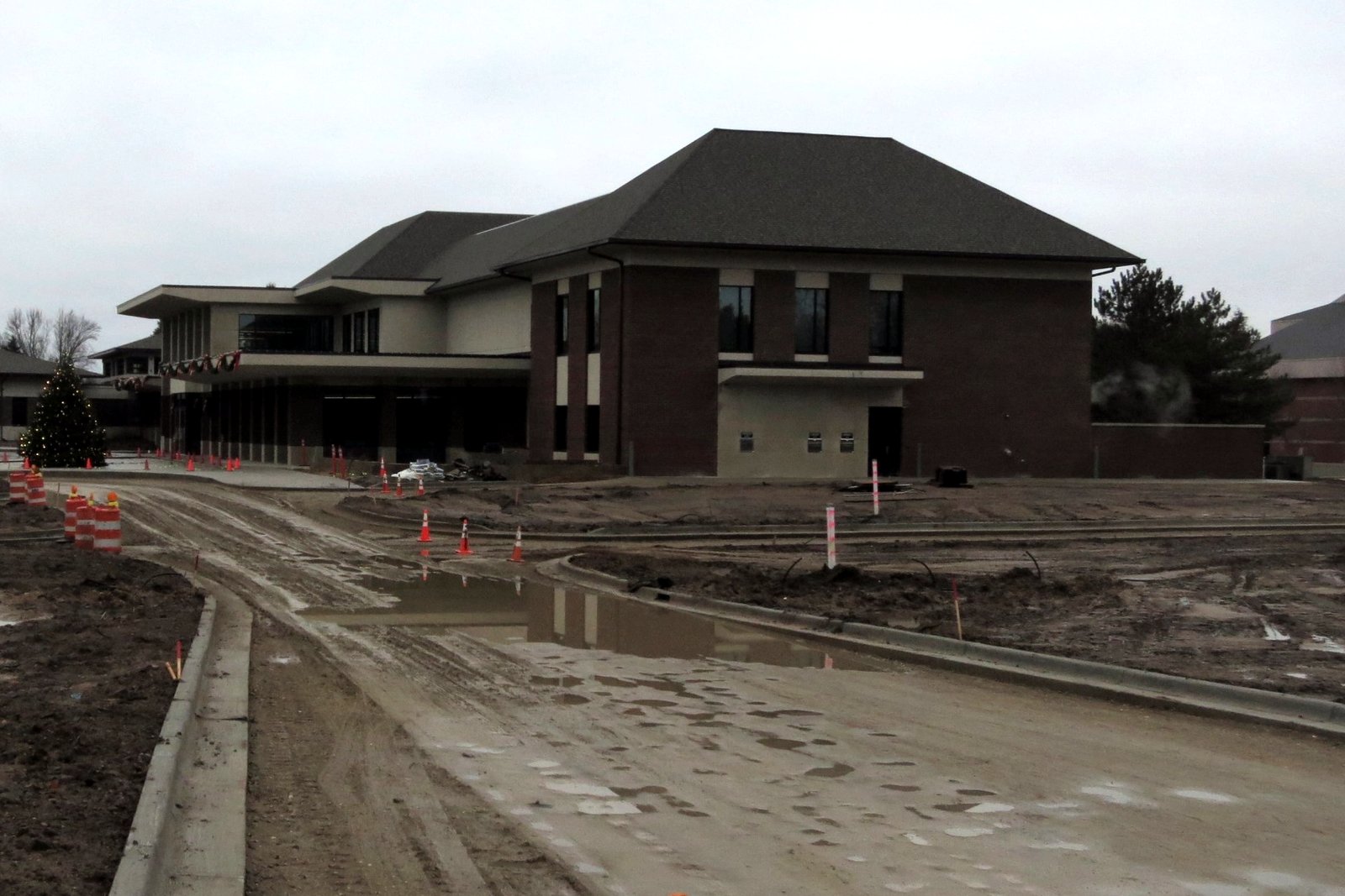 A two-story brick library building with a dark hip roof under a gray sky, with an unfinished driveway and orange construction barrels in front.