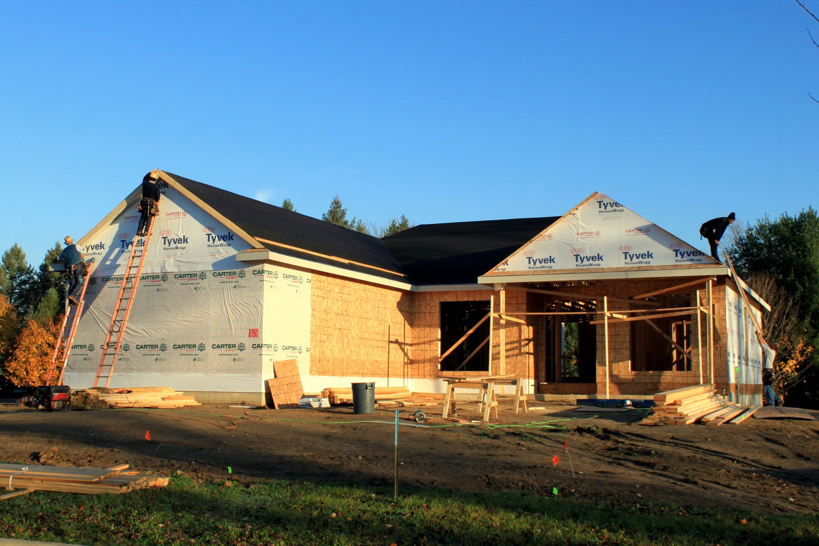 Workers on ladders and a roof finish a one-story house covered in house wrap and plywood under a clear blue sky, with lumber stacked on bare dirt in front.