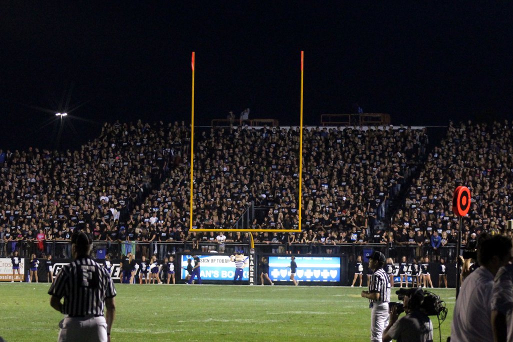 A packed grandstand of fans in dark shirts behind a yellow goalpost at night, with a referee, camera operators and sideline staff on the field in the foreground.