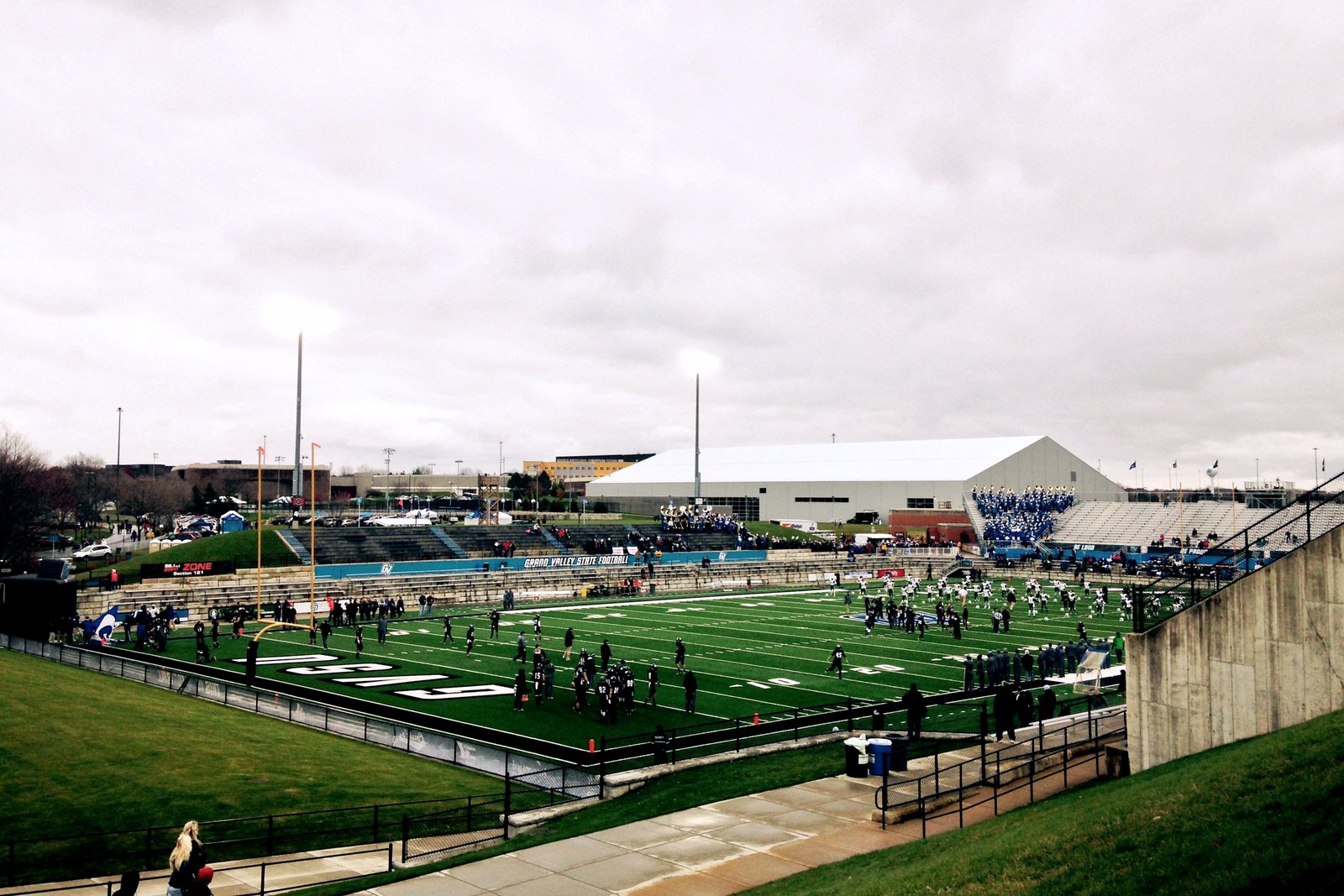 Players warm up on the green artificial turf at Lubbers Stadium under a gray overcast sky, with light towers, bleachers and a white indoor practice building beyond the field.