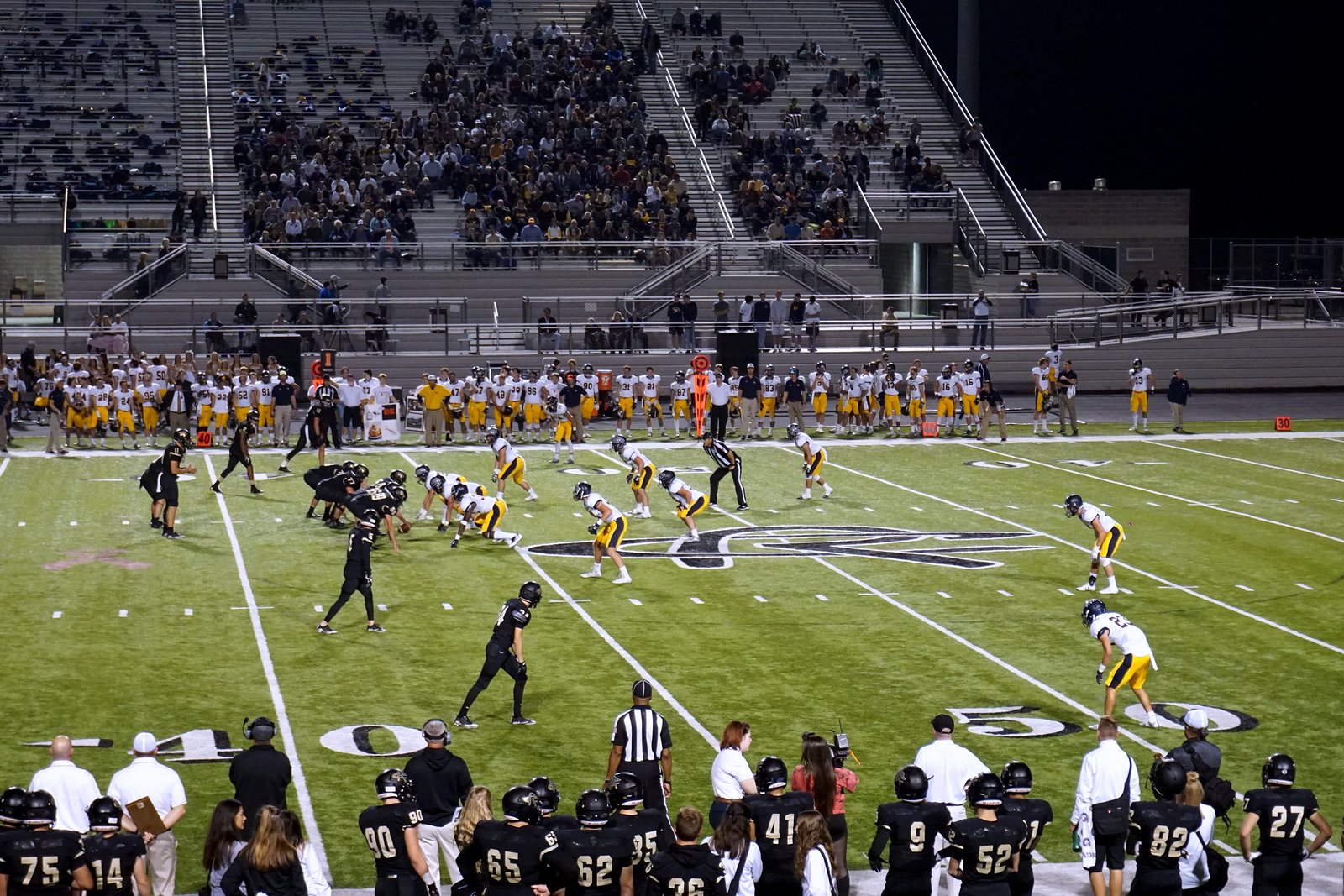High school football players run a play on a green turf field in front of a crowded grandstand, while a team in dark jerseys watches from the sideline.