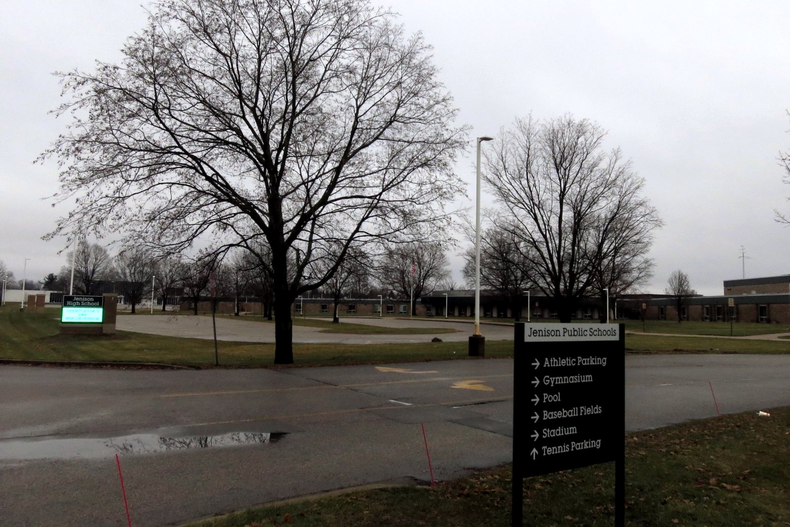 A Jenison Public Schools sign points the way to athletic parking, the gymnasium, pool, baseball fields, stadium and tennis parking on a gray day, with the school building behind it.