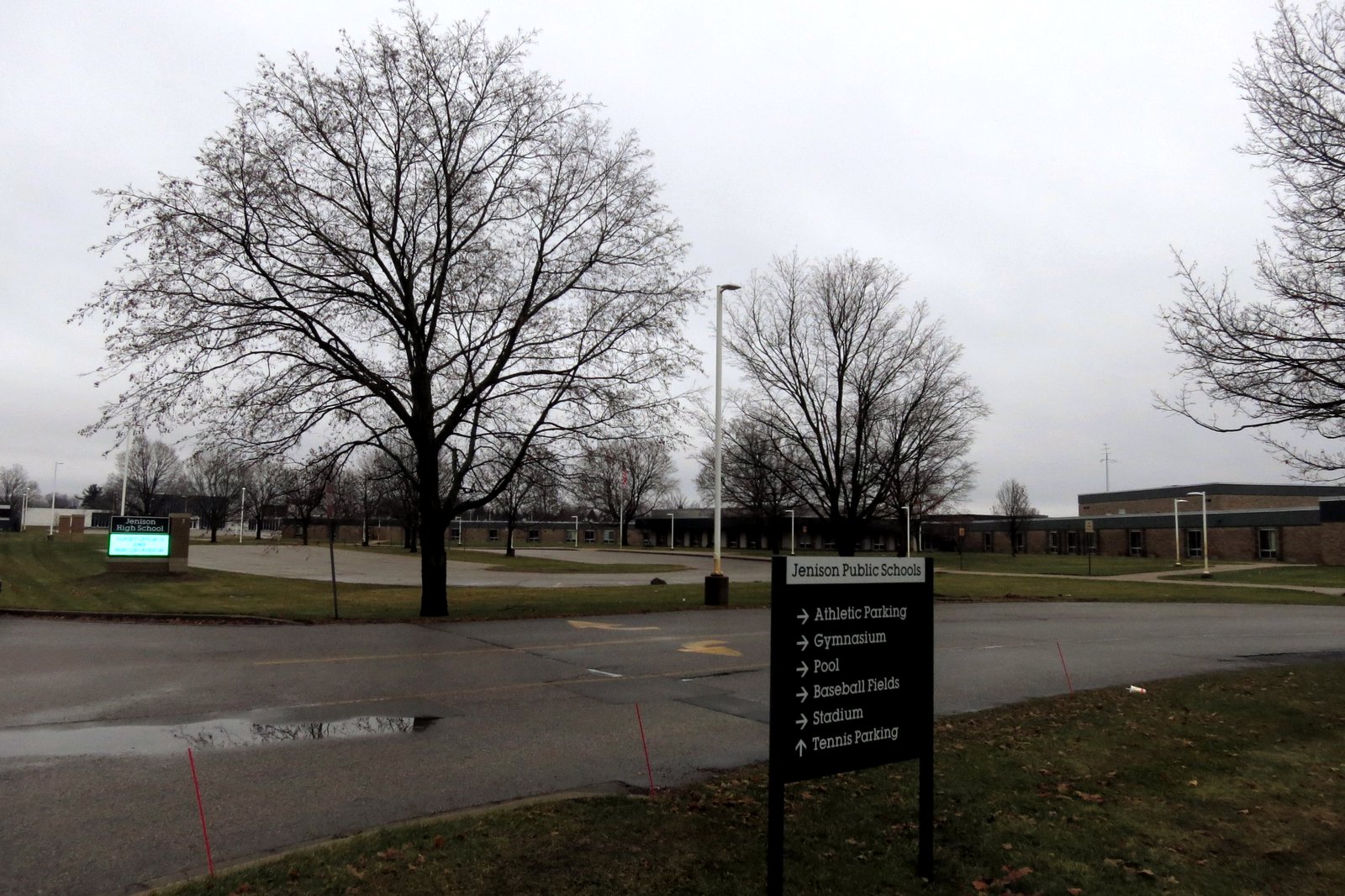 Low brick school buildings beyond a lawn and bare trees on a gray day, with a Jenison Public Schools sign pointing to the gymnasium, pool and stadium in the foreground.