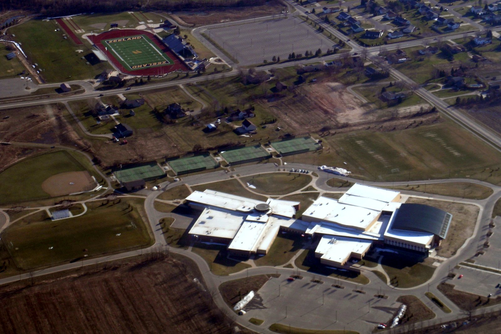 An aerial view of Zeeland West High School’s white-roofed buildings, parking lots and athletic fields, including a football stadium ringed by a red track, surrounded by farm fields and homes.