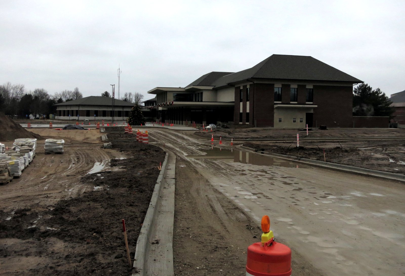 A brick building with a dark hipped roof beyond a muddy construction site, freshly poured curbs and an orange traffic barrel, under a gray sky.