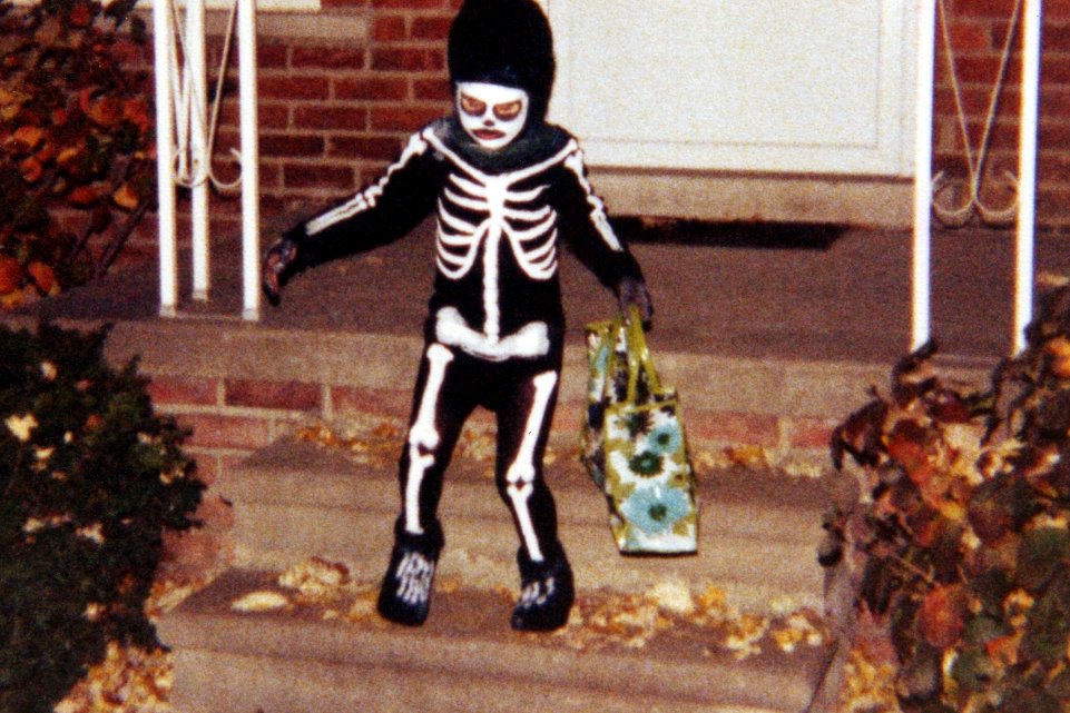 A child in a black skeleton costume stands on the brick steps of a house holding a trick-or-treat bag, with fallen leaves around the steps.