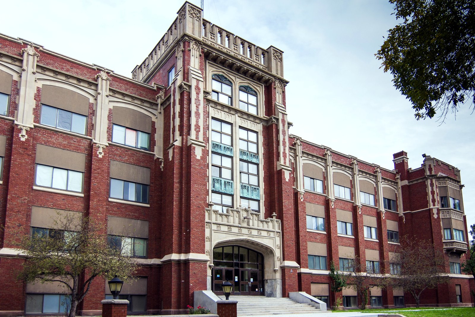 A large red-brick high school with a central tower, tall arched windows and stone trim, seen from a corner with trees and a lawn in front.