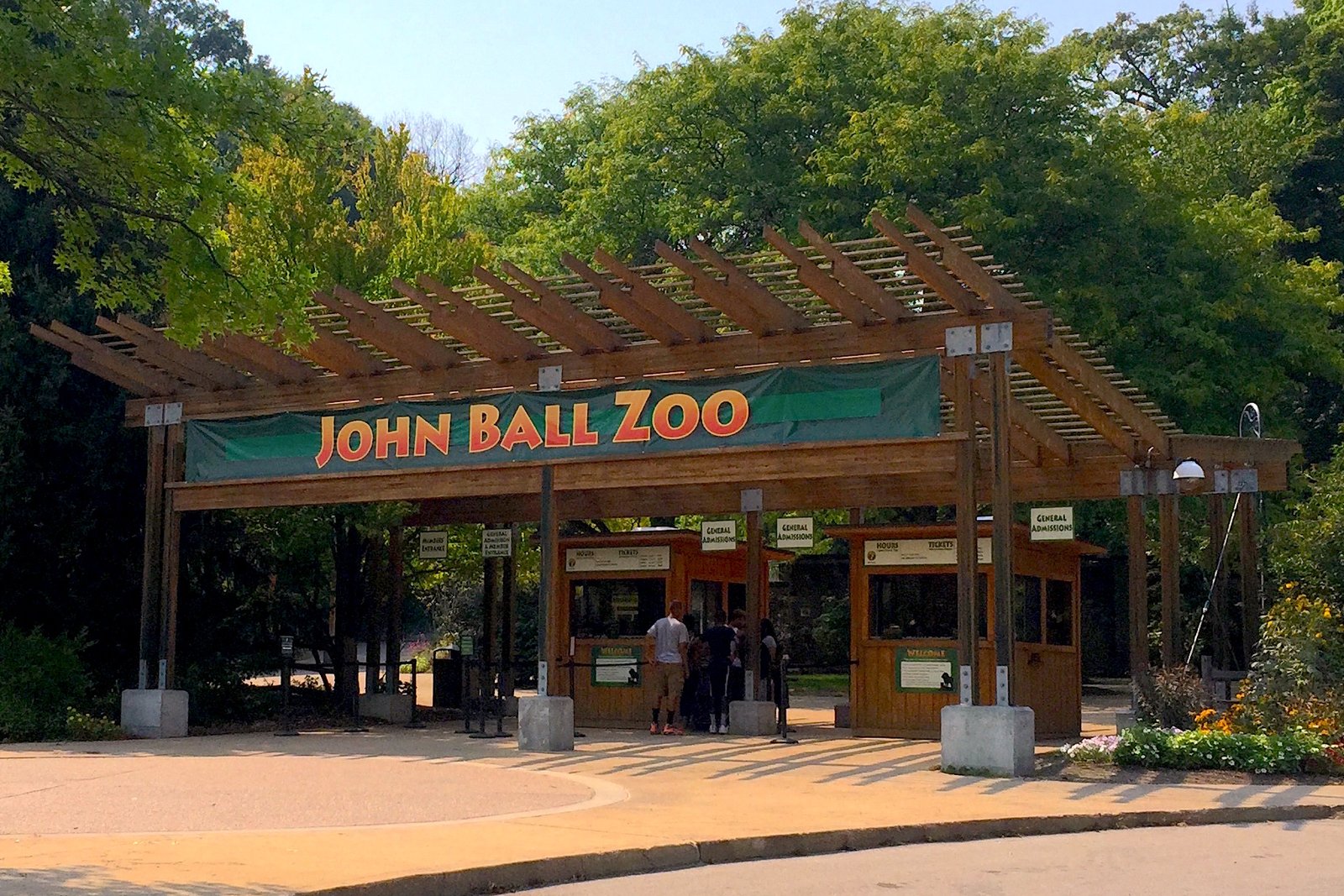 The entrance to John Ball Zoo, a timber pavilion with a green John Ball Zoo sign over the gates, with trees behind it.