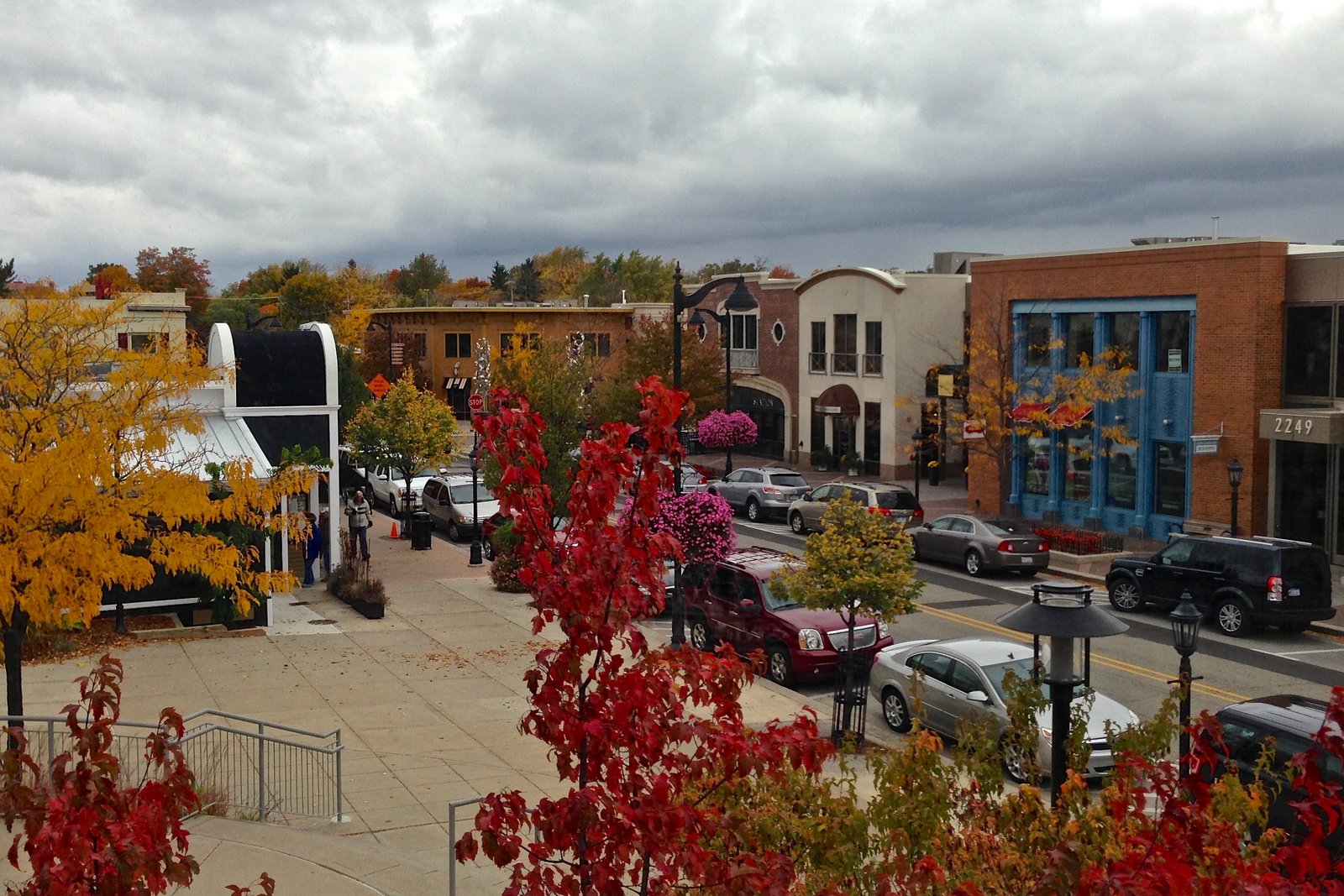 A downtown street lined with brick and stucco storefronts and parked cars under a cloudy sky, framed by trees with red and yellow fall leaves.