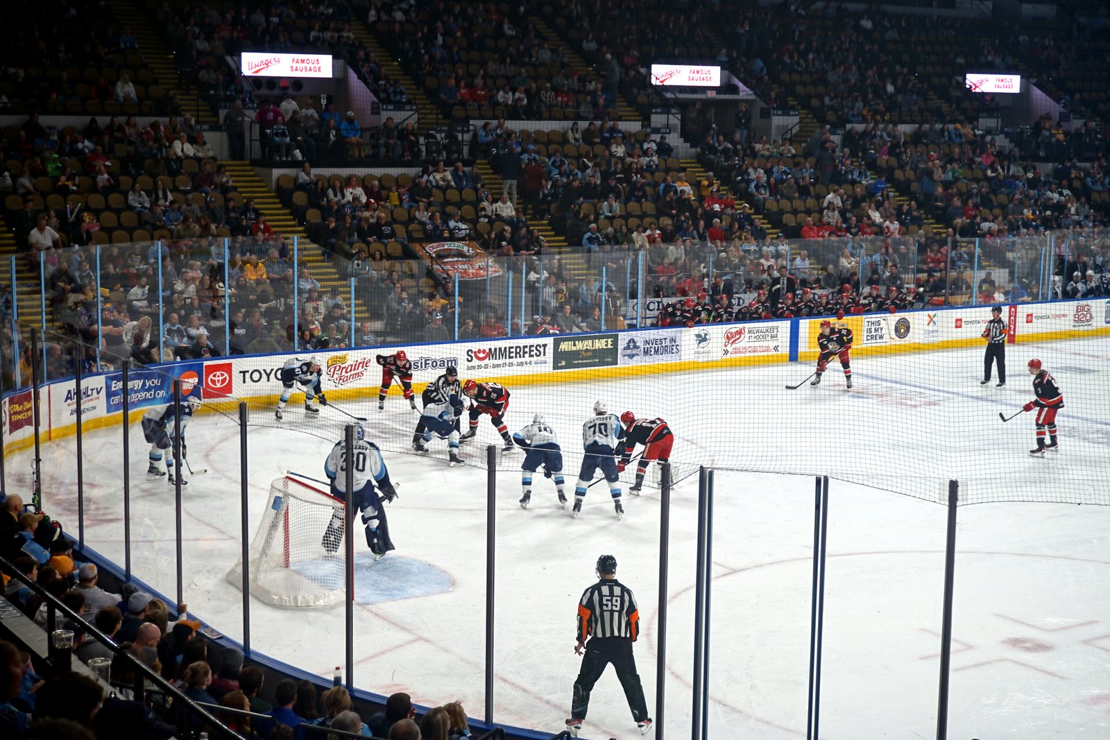 Hockey players line up for a face-off near a net as officials watch, with fans filling the lower bowl of an arena.