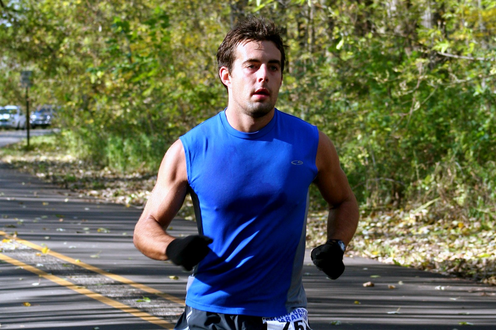 A runner in a blue sleeveless shirt and black gloves runs along a paved trail lined with autumn trees.