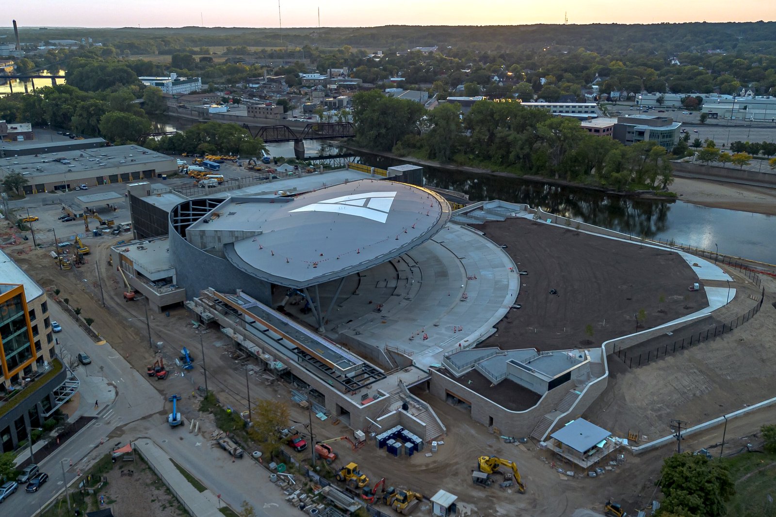 Aerial view at dusk of the Acrisure Amphitheater under construction beside the Grand River in downtown Grand Rapids, with its curved white roof and open lawn seating.