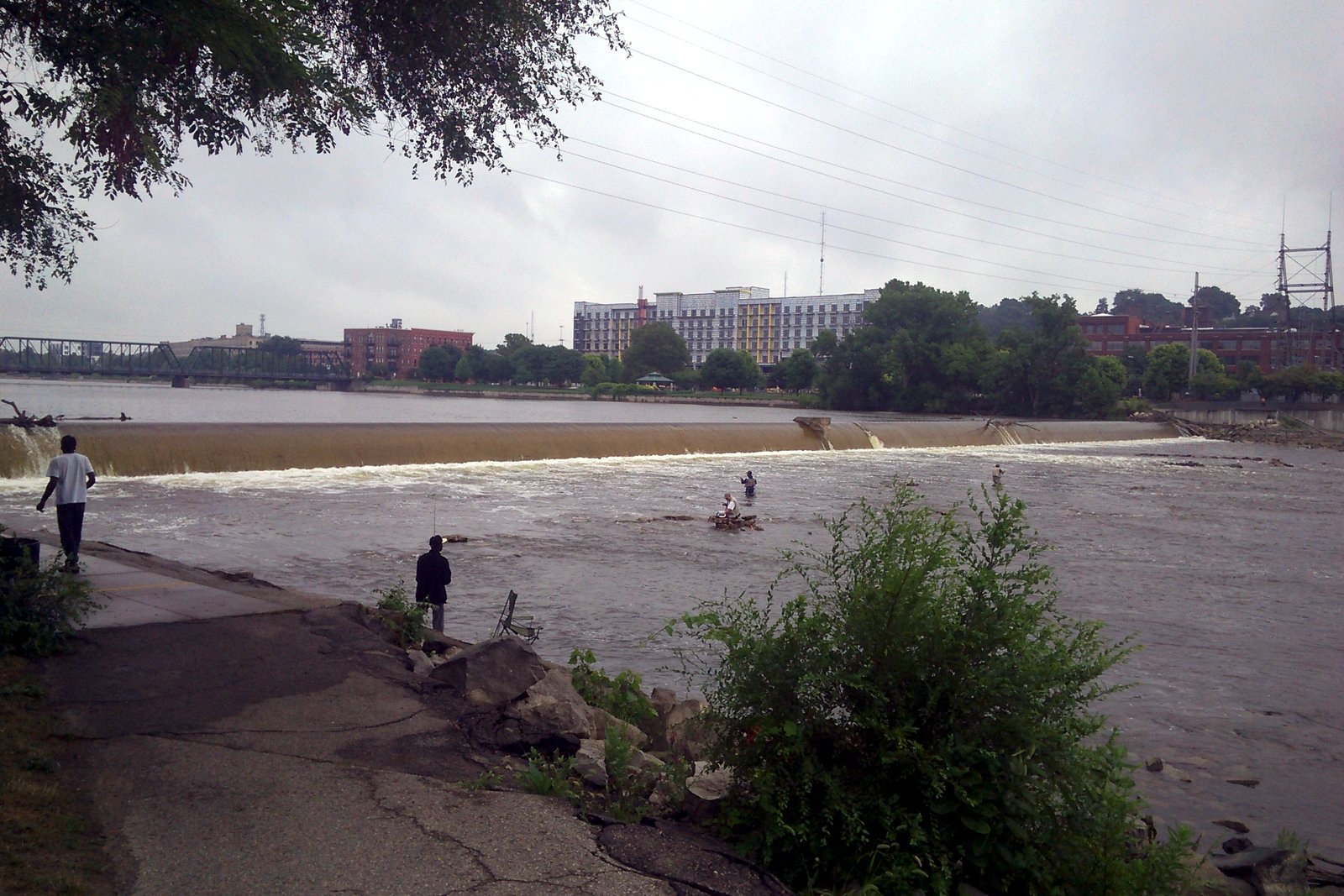 Anglers stand on a riverside walkway and wade in the Grand River below a low dam that spans the river, with a truss bridge and brick and mid-rise buildings on the far bank.