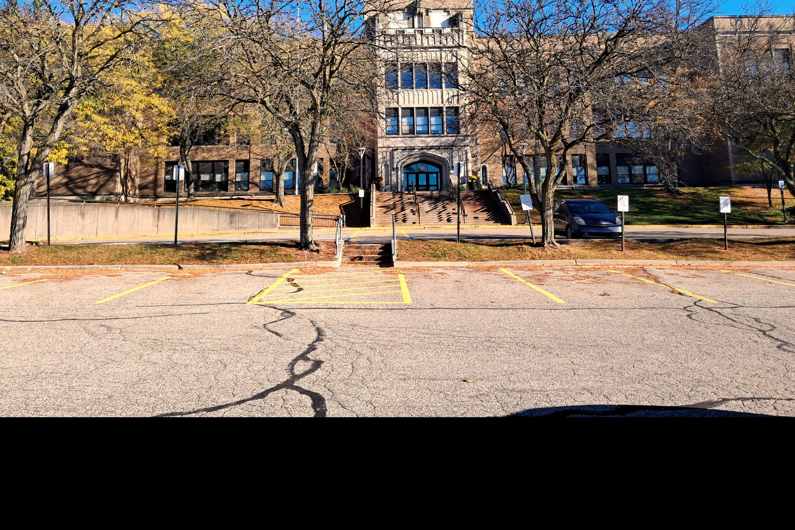 A multistory brick school building behind bare trees and a parking lot on a sunny autumn day.