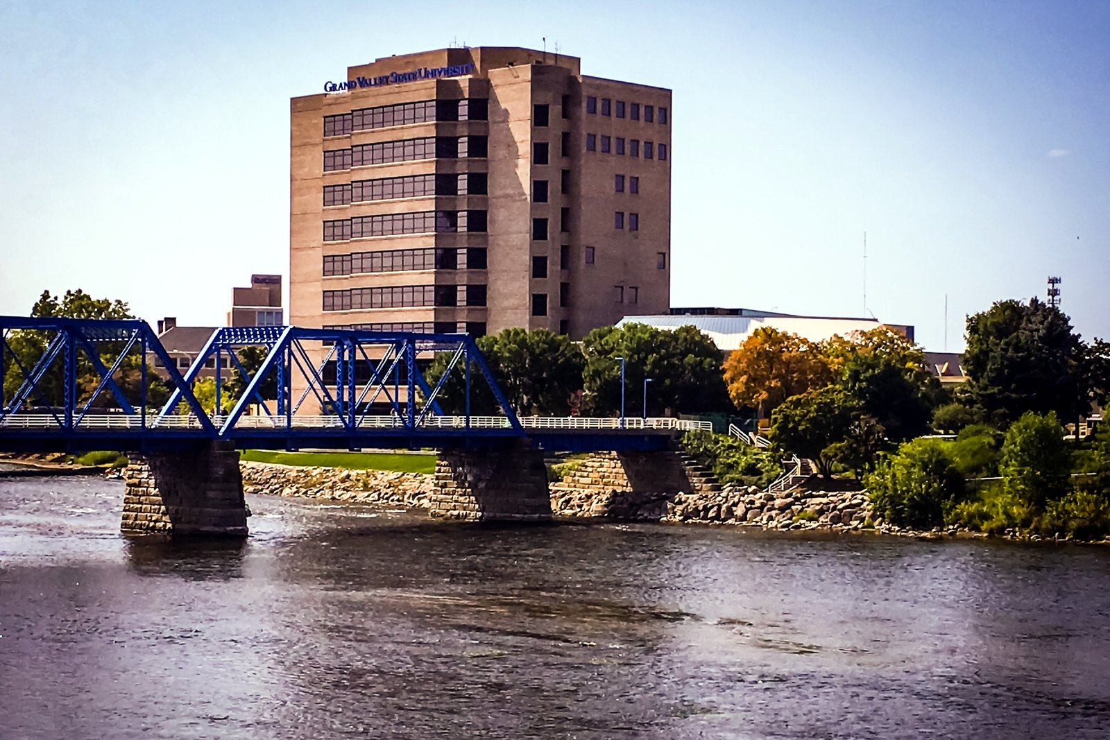 A blue steel truss bridge crosses a wide river toward a tall brick office building and trees in early fall color under a blue sky.