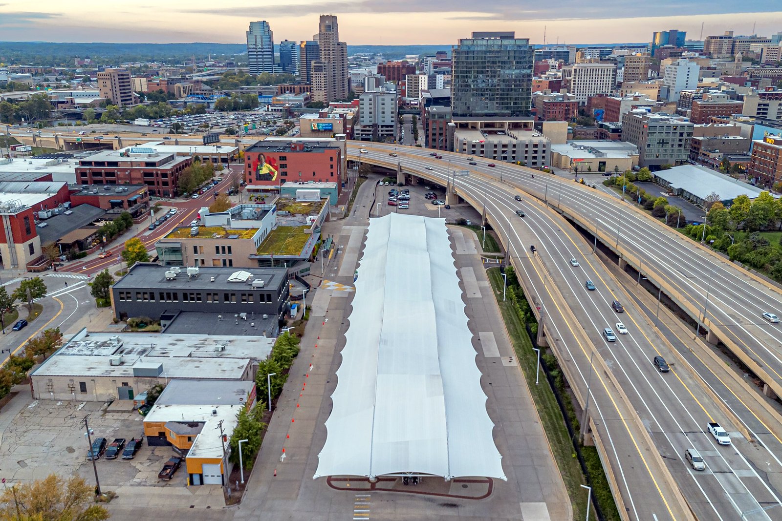 Aerial view of a curving elevated freeway passing beside downtown Grand Rapids buildings and a covered parking area at dusk.