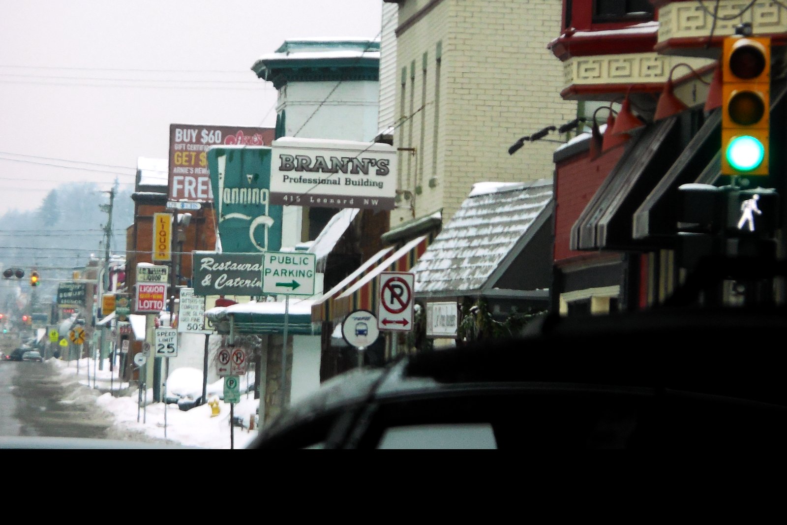 A snowy commercial street lined with older brick and painted storefronts, with a sign reading Brann’s Professional Building, 415 Leonard NW, and a green traffic light in the foreground.