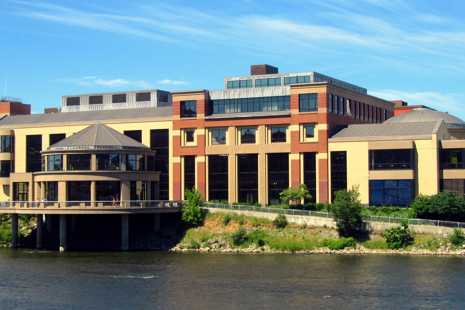 The Grand Rapids Public Museum, a tan and red brick building with a round glass pavilion, seen across the Grand River on a sunny day.