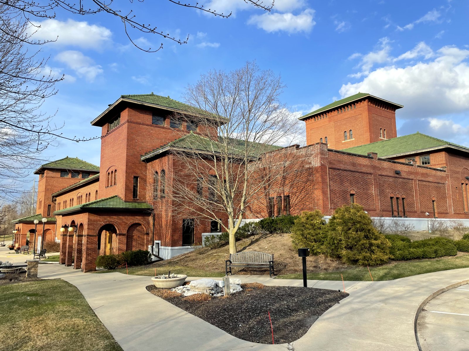 A long red-brick Romanesque building with green-trimmed hip roofs and arched entries sits behind a curving concrete walk and bare trees under a blue sky.