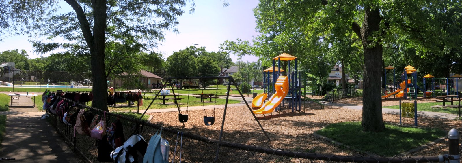 A shaded city park with tall trees, a swing set and a playground with orange slides and yellow roofs, a fence hung with backpacks in the foreground and a pool and low brick building in the distance.