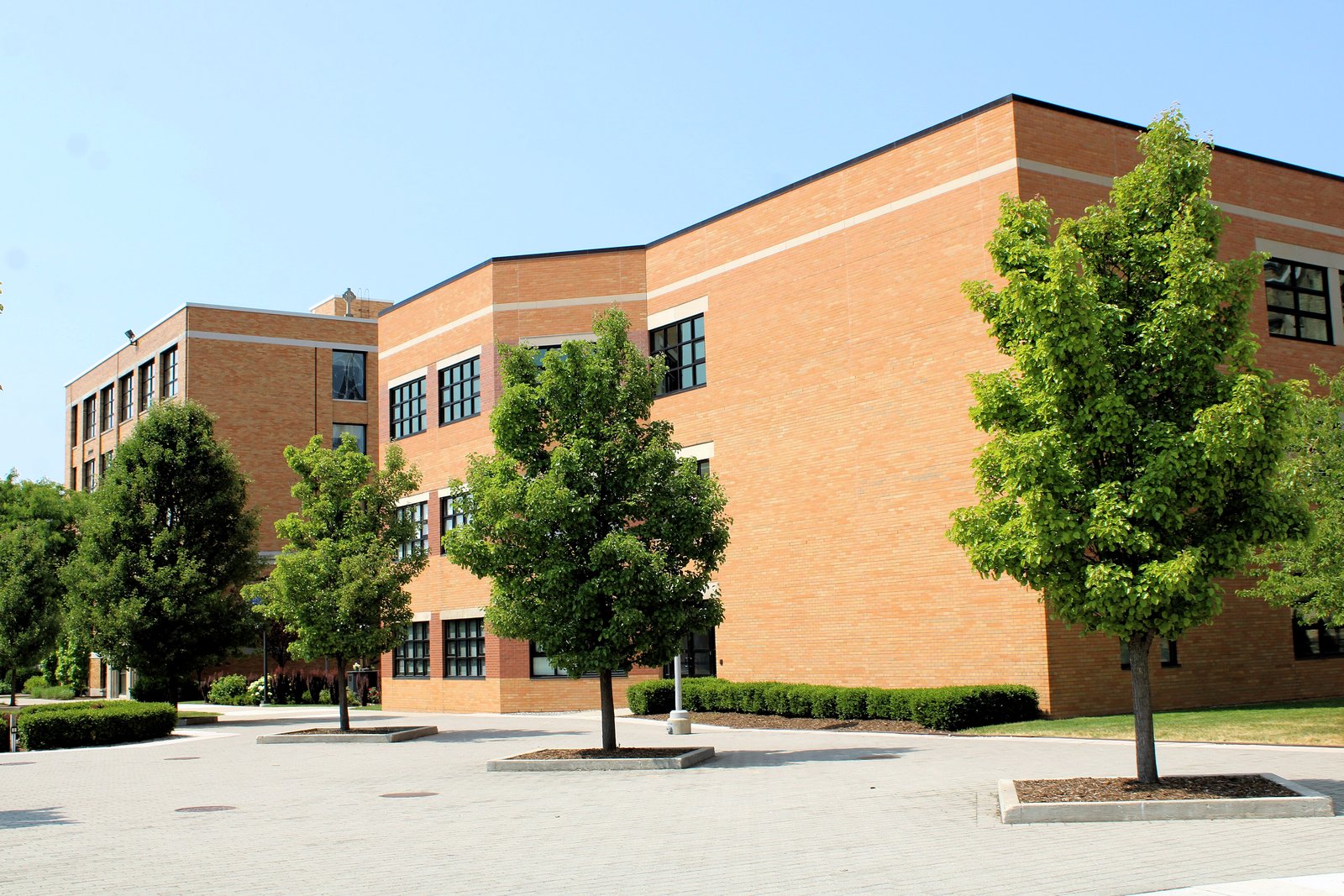 A two-story tan brick school building with tall windows and young trees along a paved walkway on a sunny day.