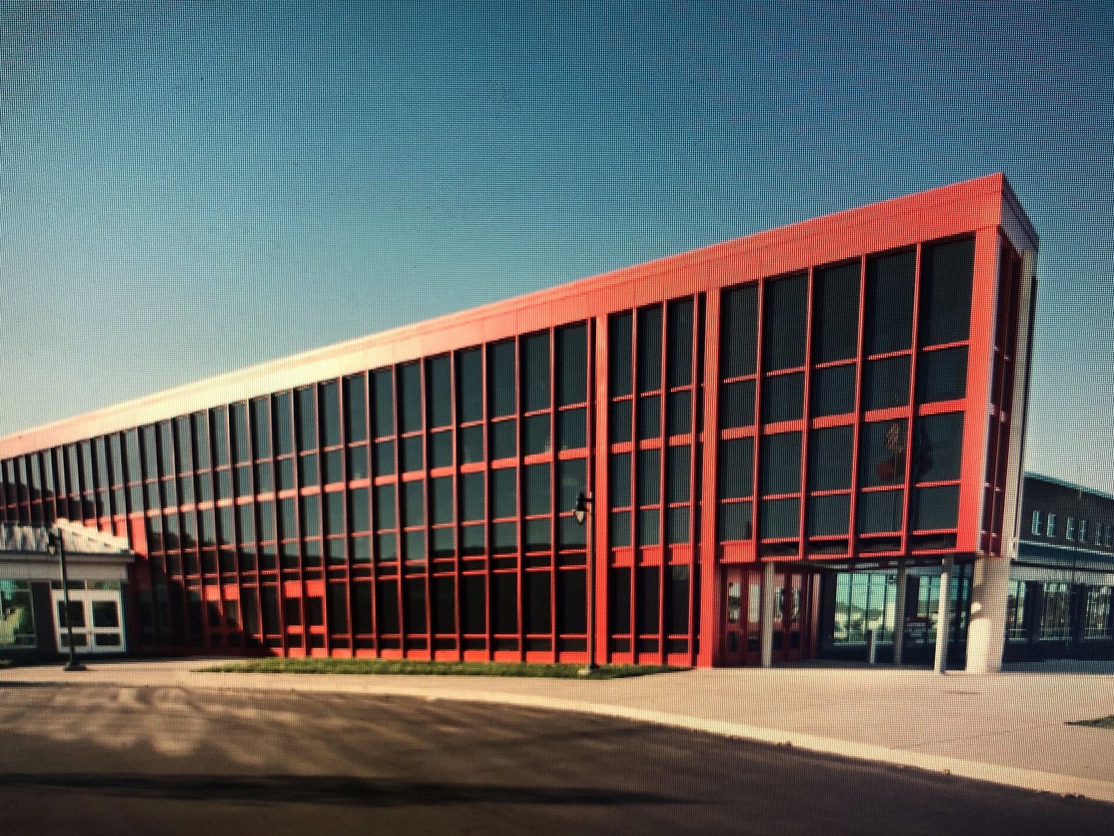 A long, low school building with a bright red frame and dark glass windows rising to a tall corner at the right, under a clear blue sky.