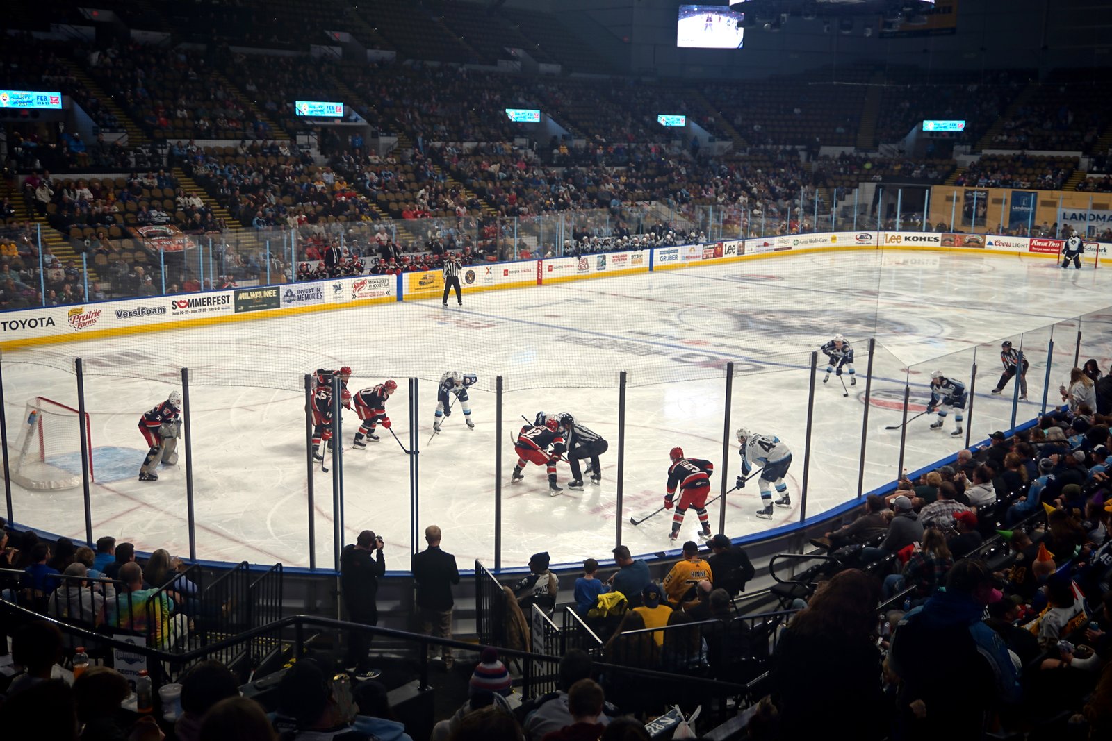 Hockey players line up for a face-off on a white rink in front of a crowd, seen from high in the stands.